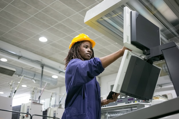 Focused confident female factory worker operating industrial machine, touching control board, using tablet