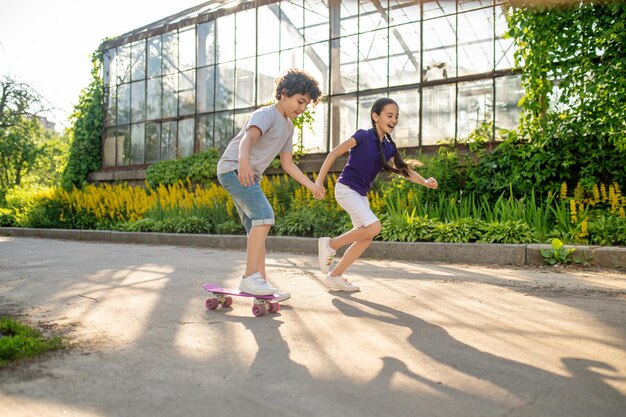 Focused curlyhaired boy learning to ride the skateboard accompanied by a cheerful cute girl