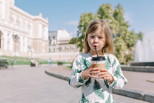 Focused cute little girl in summer dress drinking cocoa outside in sunlight