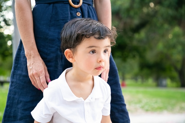 Focused excited little black haired boy standing by mom outdoors and staring away. Medium shot. Childhood concept