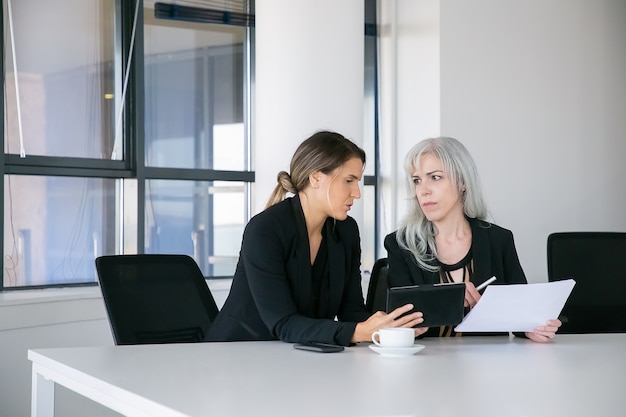 Focused female colleagues discussing and analyzing reports. Two professionals sitting together, holding documents, using tablet and talking. Teamwork concept