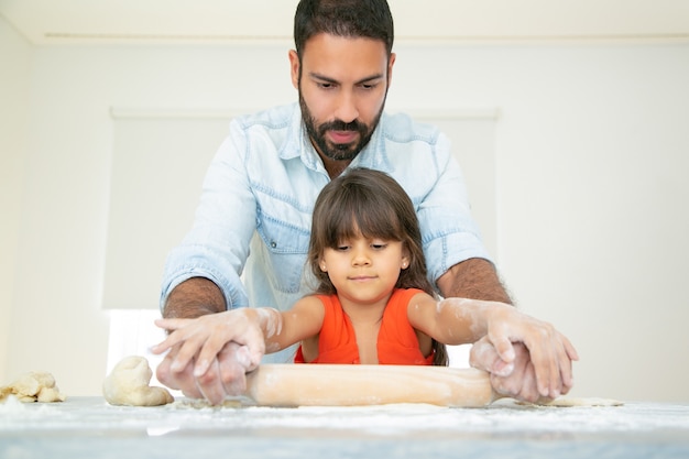 Focused girl and her dad kneading and rolling dough on kitchen table with flour messy.