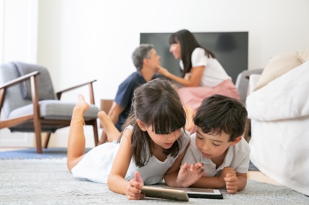 Focused little kids lying on floor in living room and using digital devices with learning apps while parents kissing