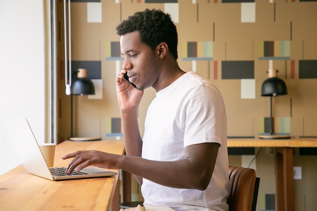 Focused young entrepreneur working at laptop and talking on mobile phone in co-working space