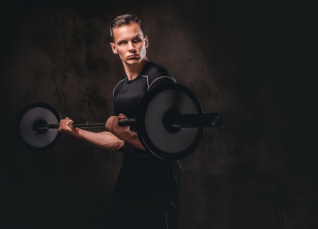 Focused young sportsman holding a barbell and doing exercise on biceps. Isolated on a dark background.