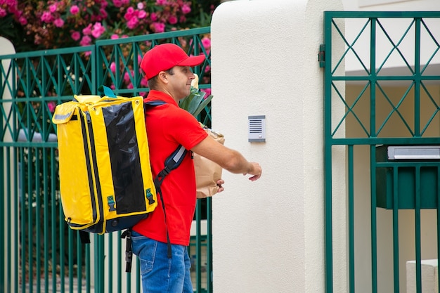 Food courier in uniform with isothermal backpack and paper package ringing doorbell. Shipping or delivery service concept