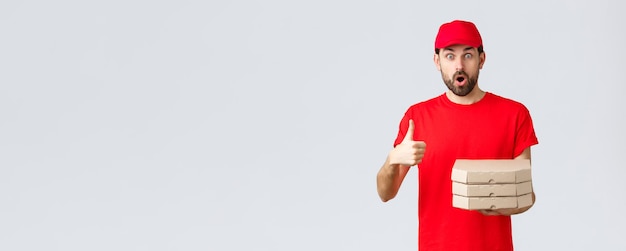 Free photo food delivery, quarantine, stay home and order online concept. surprised and amazed bearded courier in red t-shirt, cap gasping impressed, show thumb-up, bring order pizza, grey background.