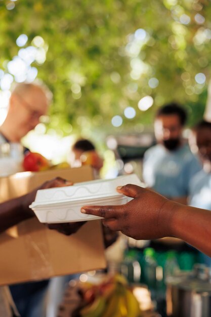 At food drive, volunteer with african american ethnicity serves a poor, hungry person a warm meal. Close-up of a less privileged needy individual receiving free food from charity worker.