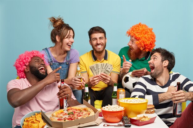 Football fans, happiness and fun concept. Overjoyed friend glad have success on football bet, win lump sum of money, hold dollars, eat tasty snack, sit around table, laugh loudly, isolated on blue