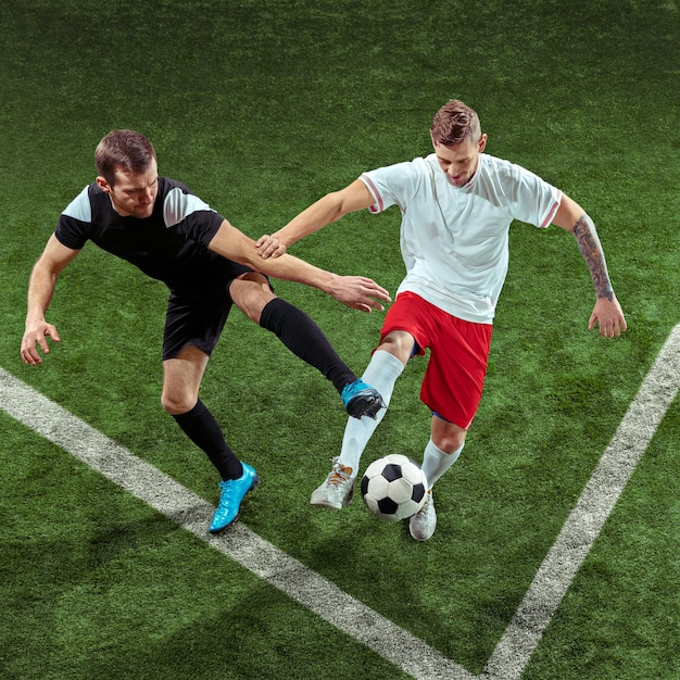 Football player tackling for ball over green grass wall. Professional male soccer players in motion at stadium. Fit jumping men in action, jump, movement at game.