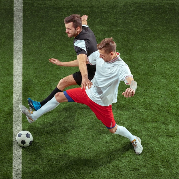 Football player tackling for ball over green grass wall. Professional male soccer players in motion at stadium. Fit jumping men in action, jump, movement at game.