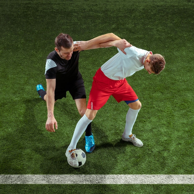 Football player tackling for ball over green grass wall. Professional male soccer players in motion at stadium. Fit jumping men in action, jump, movement at game.