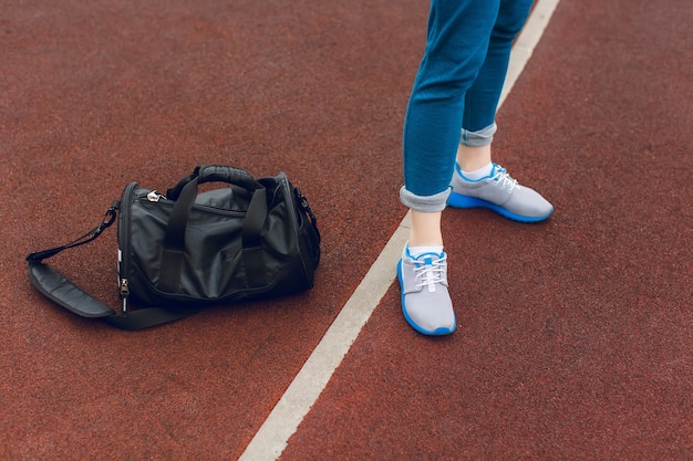 The foots in gray sneakers are standing near white line on the staduim. There is a black sport bag near.