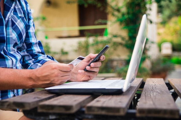 Free photo forty years old caucasian man looking at credit card while working on laptop computer on garden terrace during sunny summer day. modern lifestyle - countryside weekend and shopping online concept.