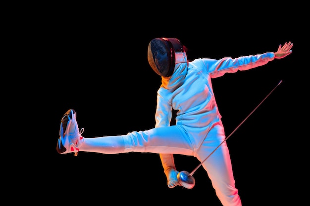 Forward. Teen girl in fencing costume with sword in hand isolated on black wall, neon light. Young model practicing and training in motion, action. Copyspace. Sport, youth, healthy lifestyle.