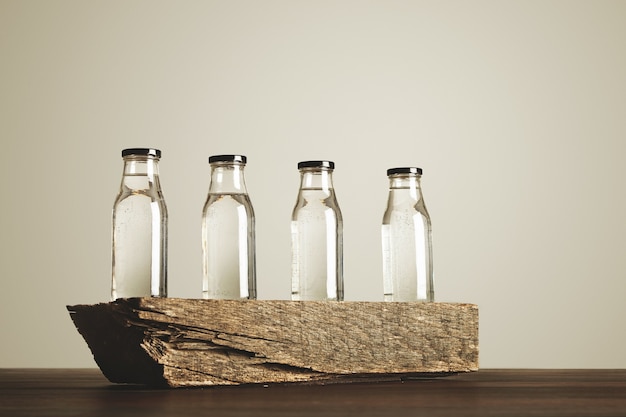 Free photo four clear transparent glass bottles with black caps filled with pure drinking water presented on wooden brick, isolated on white