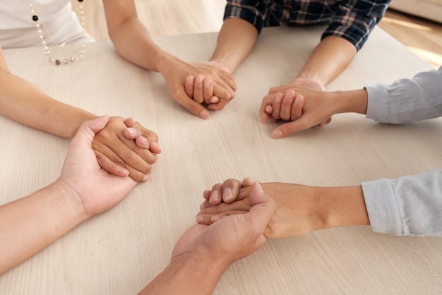 Four unrecognizable people sitting around table and holding each other's hands in middle