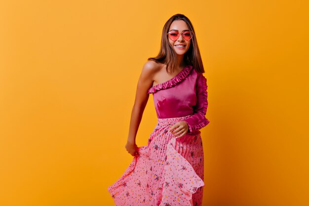 Frail girl in stylish pleated skirt and heart-shaped glasses moves her outfit. Stylish portrait in full growth on orange wall