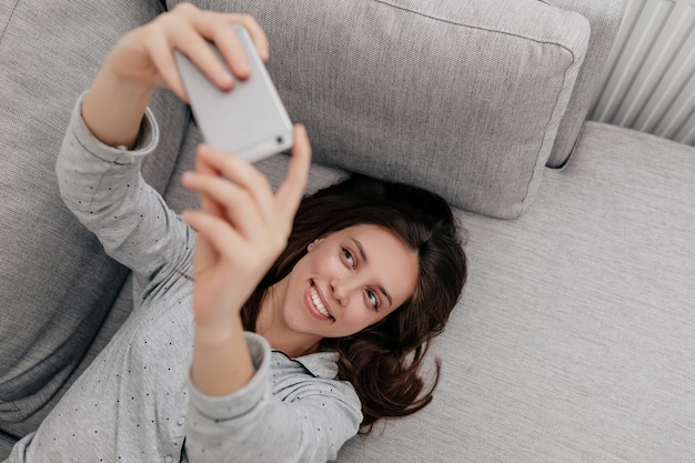 Frame above of lovely attractive smiling woman with dark hair wearing pajamas lying on the coach and making selfie with smartphone.