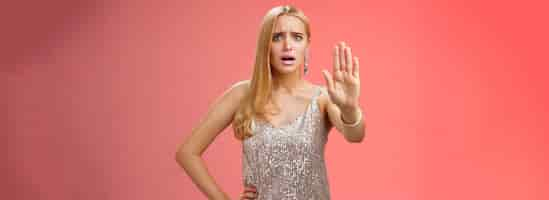 Free photo freaked out displeased bothered insecure blond woman in silver glittering dress extend palm stop enough prohibition rejection gesture bothered pissed annoying clingy man nightclub red background