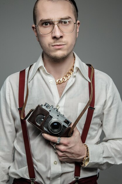 Freaky guy in sunglasses and white shirt holds film camera over grey background.