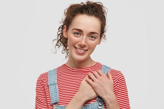 Freckled young curly female keeps hands on heart, expresses good attitude, dressed casually, shows natural beauty, being in high spirt, isolated over white wall. Friendly smiling girl.