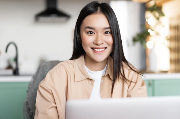 Freelance asian girl sitting at home and working with laptop smiling at camera young woman using com...