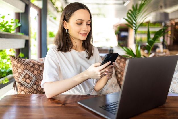 Freelance young woman sitting in the cafeteria with laptop and drinking coffee while use her phone