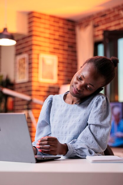 Freelancer answering phone call to discuss work plan with clients in modern home office with beautiful warm sunset light. African american woman chatting with company colleagues on smartphone