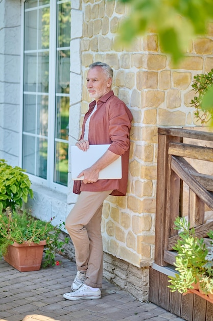 Freelancer. Mature man in casual clothes with a laptop near a brick wall