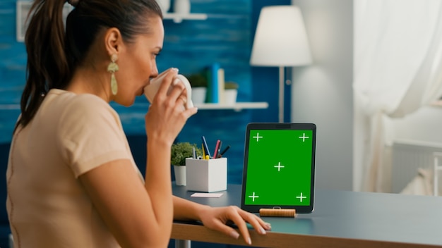 Free Photo freelancer woman holding cup of coffee looking at tablet computer with mock up green screen chroma key sitting on desk table. caucasian female browsing on isolated device from home office room