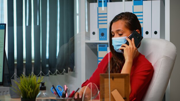 Freelancer working and talking on phone sitting at workplace wearing protection face mask during coronavirul pandemic. Woman chatting with remotely team speaking on smartphone in front of computer