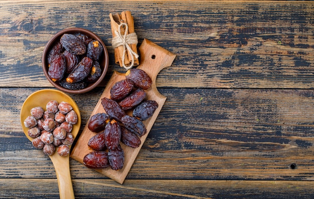 Fresh dates in bowl and cutting board with nuts in wooden spoon and cinnamon sticks top view on wooden background