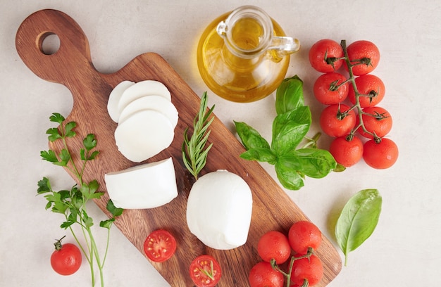 Fresh mozzarella cheese, Soft italian cheeses, tomato and basil, olives oil and rosemary on wooden serving board over light wooden surface. Healthy food. Top view. Flat lay.