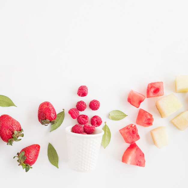 Fresh raspberries spilled front glass with strawberry; watermelon and pineapple isolated on white background