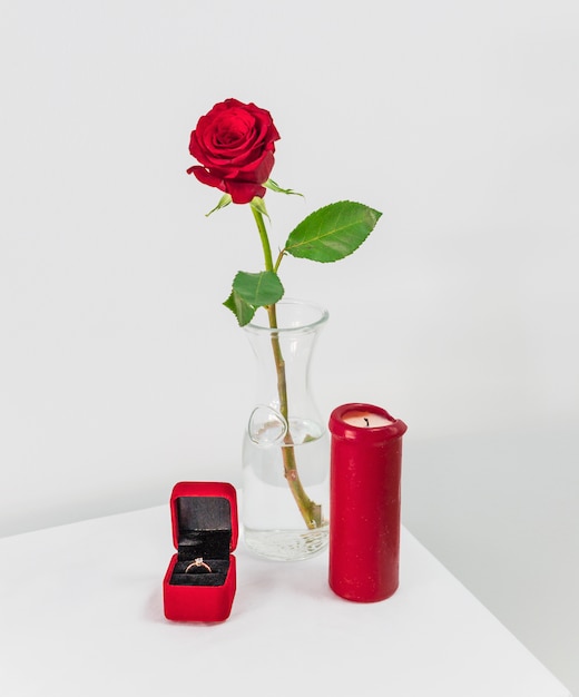 Fresh red rose in vase and present box with ring near candle on table