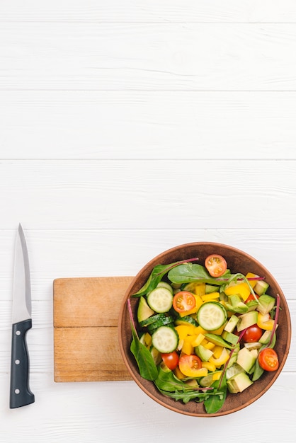 Free photo fresh salad bowl on chopping board with sharp knife on white table