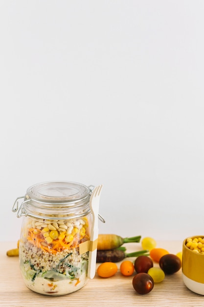 Free photo fresh spring salad in closed jar with white fork and vegetables on wooden table