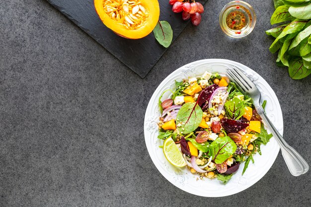 Fresh vegetable salad with beetroot, arugula, red onion, sorrel, chickpeas, pumpkin and grapes in a white plate on a black table. Top view