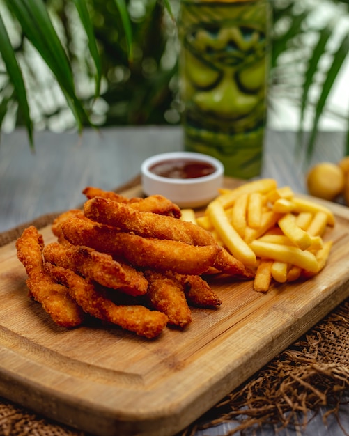 Fried breaded chicken breast strips with ketchup and french fries on a wooden board