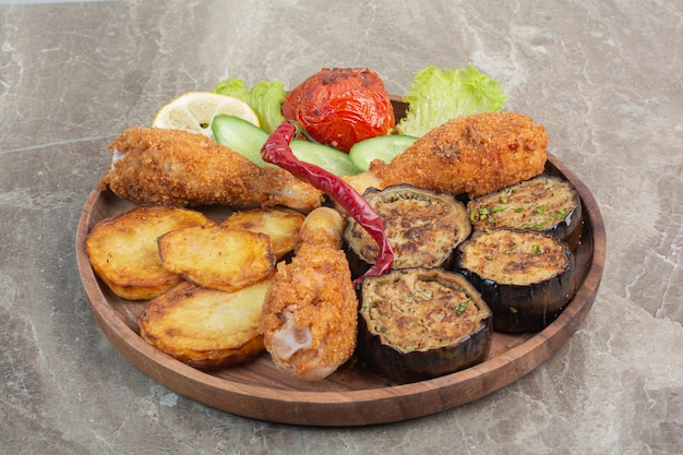 Fried chicken legs with potato and eggplant on wooden board.