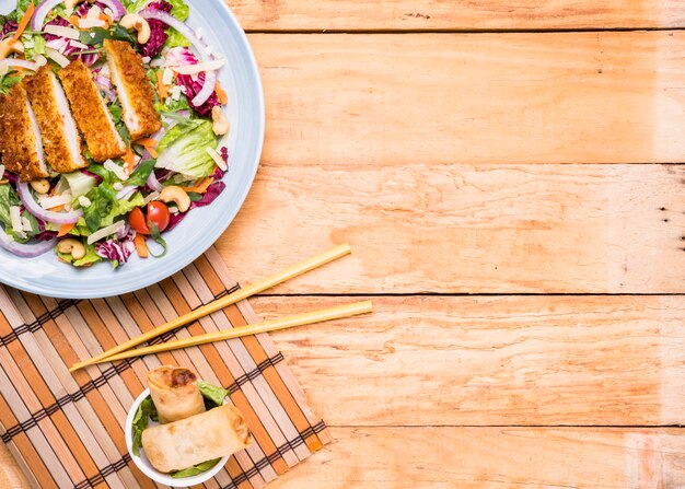 Free photo fried fillet salad with spring rolls in ceramic bowl and chopsticks against wooden table