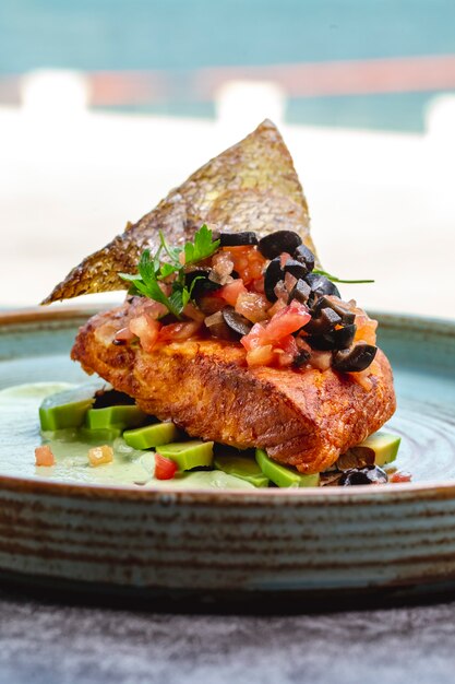 Fried fish fillet placed on avocado slices and sauce topped with tomato olive and fish skin