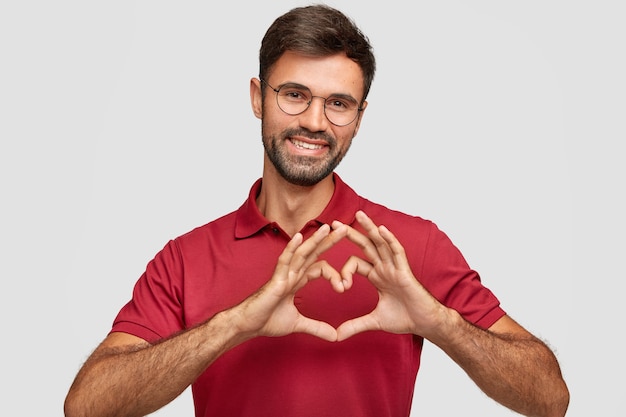 Free photo friendly attractive bearded male makes heart gesture, smiles pleasantly, wears spectacles and red t-shirt, expresses love