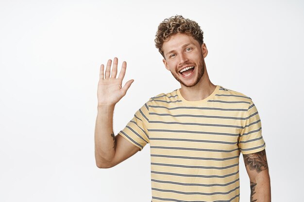 Friendly blond guy saying hello waving hand and smiling at camera standing over white background