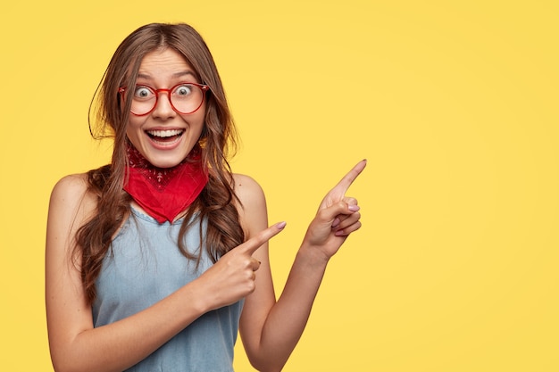 Friendly carefree female shop assistant points right, has board smile, advertises new outfit with big discounts, wears transparent glasses, models against yellow  wall with copy space for slogan