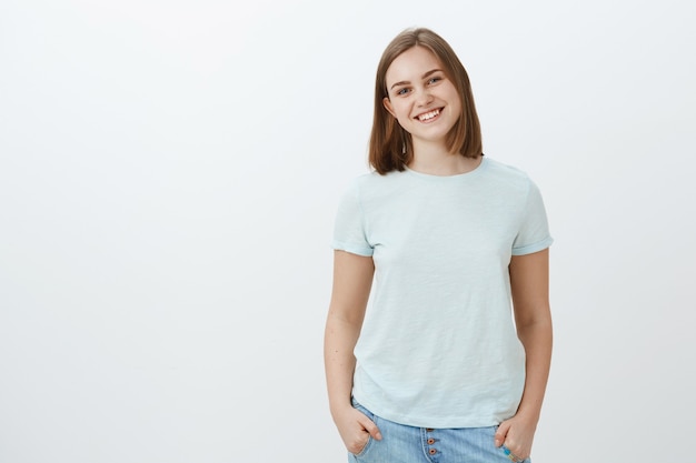 Friendly cute white girl with short haircut and lovely flapped ear smiling joyfully holding hands in pockets and gazing with joyful entertained look posing over white wall