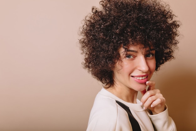 Friendly cute woman with ringlets and nude make up smiles at camera and enjoys good mood on beige