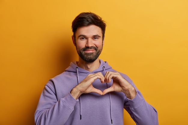 Friendly looking bearded guy shapes heart gesture, sends love, charity and volunteer, wears purple sweatshirt, poses over yellow wall, demonstrates affection