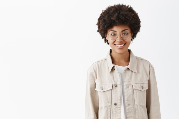 Free photo friendly-looking casual boyish dark-skinned woman with afro hairstyle in glasses and trendy shirt smiling broadly and standing over gray wall with carefree and relaxed look